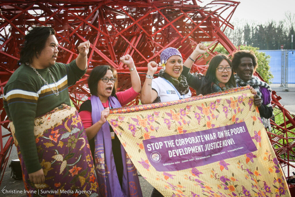 Five people stand together holding a colorful banner that reads "STOP THE CORPORATE WAR ON THE POOR! DEVELOPMENT JUSTICE NOW!" with raised fists in front of a red metal structure.