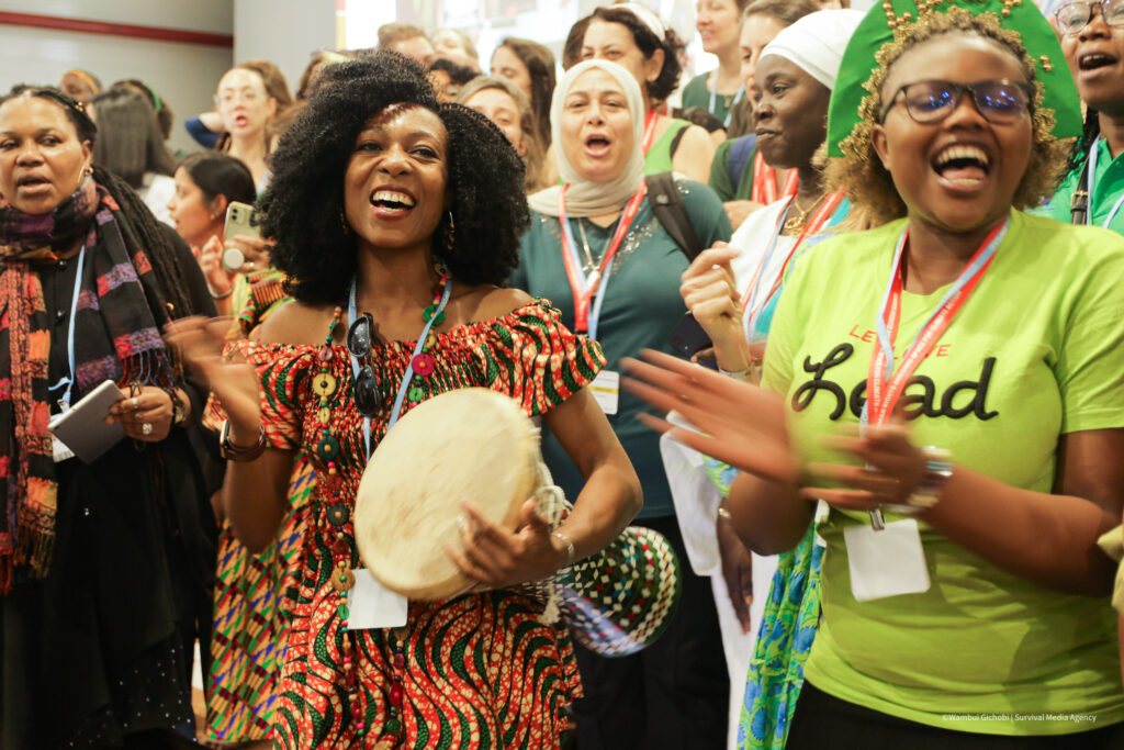 A diverse group of people joyfully singing and clapping, with one person playing a drum and another wearing a green shirt.