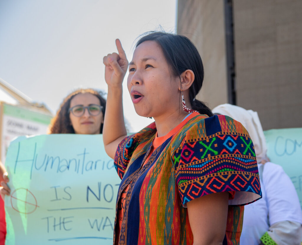 A woman in a colorful patterned outfit speaks passionately with her finger raised, standing in front of people holding a blue sign with text.