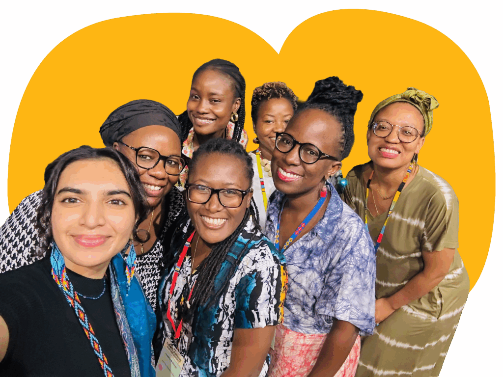 A group of women in vibrant clothing poses for a selfie at a nighttime outdoor event, with greenery and modern architecture in the background.