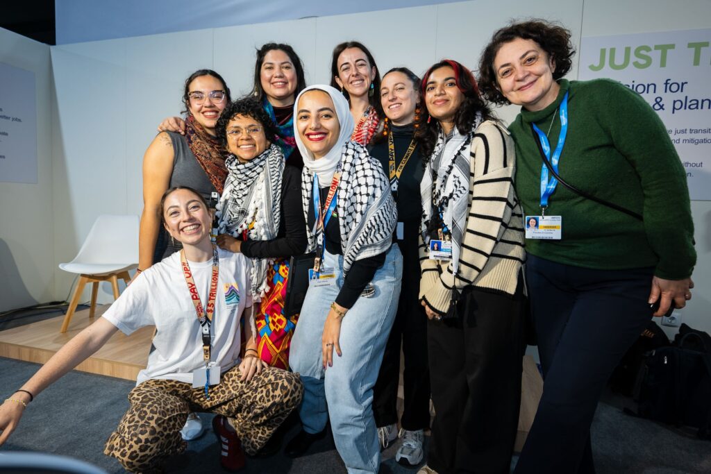 A group of nine people stands together indoors, smiling at the camera. Some wear lanyards and black-and-white kefiyahs also used as hijab. A stage and chairs are visible in the background.